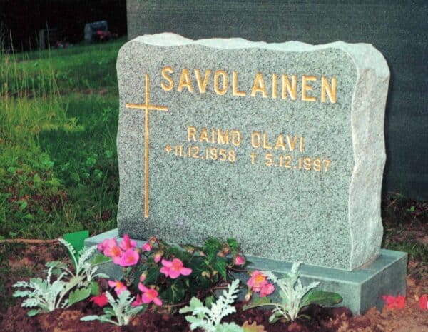 Kuvass on hautakivi Savolainen, joka on vaaleanharmaa väritykseltään ja muodoltaan suorakulmainen, mutta reunoissa on laineita. Teksti hautakivessä ja sen vasemmalla puolella oleva risti tulevat molemmat kullan värisinä.